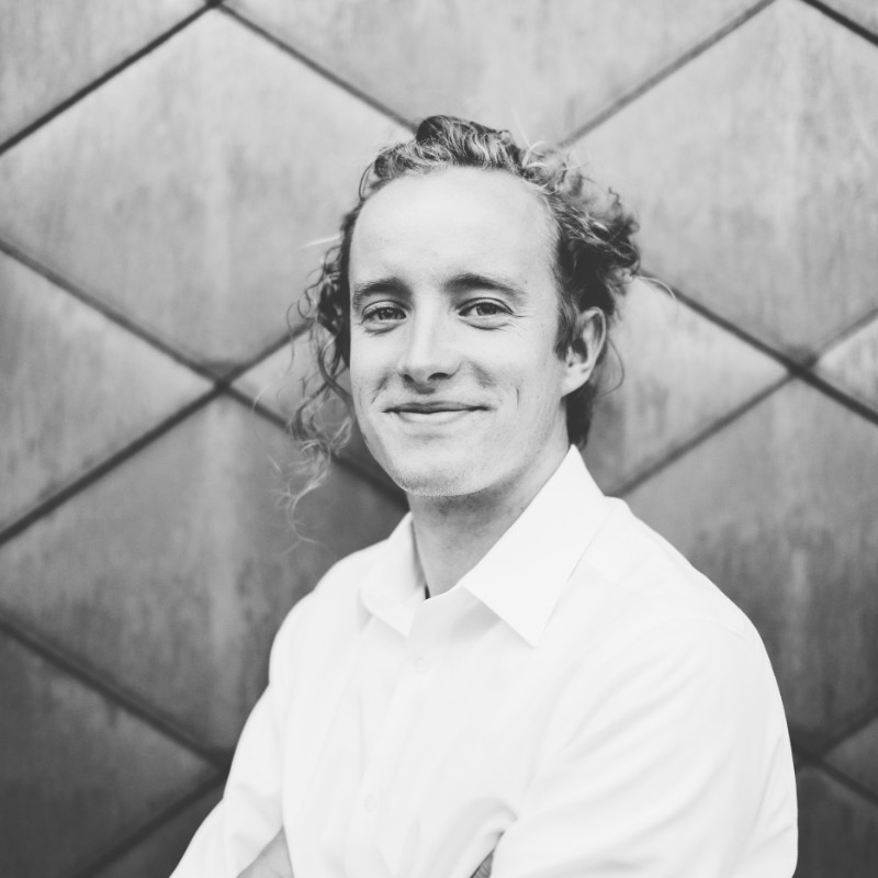 Black-and-white portrait of a smiling person in a white collared shirt against a geometric paneled wall.